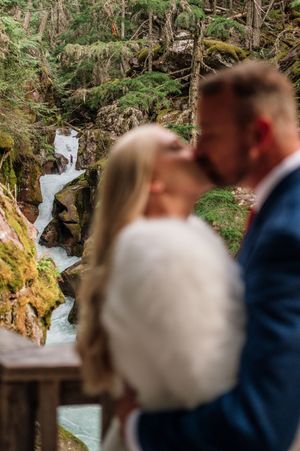 Blurred bride and groom share romantic kiss in front of turquoise waterfall cascading through mossy forest rocks.