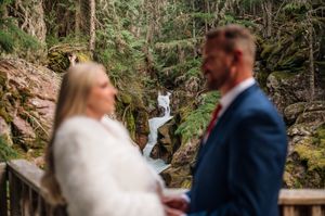Bride and groom hold hands near a waterfall in a lush forest during their outdoor wedding ceremony.