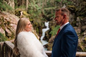 Bride in white fur wrap and groom in navy suit sharing intimate moment by waterfall in forest setting.