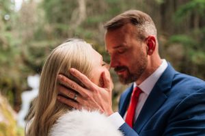 Groom tenderly cups brides face during intimate wedding moment outdoors in forest setting.