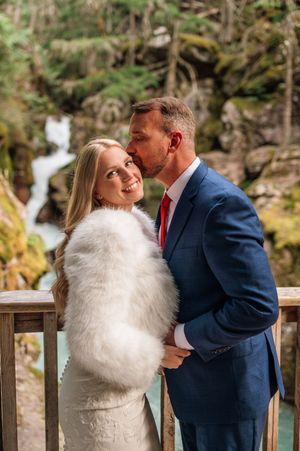 Groom in navy suit kisses bride's cheek as she smiles on wooden bridge near mossy waterfall.