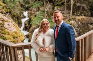 Bride and groom pose together on wooden bridge with waterfall and rocky backdrop.