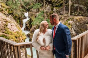 Couple in wedding attire smiling on wooden overlook with waterfall and mossy rocks behind.