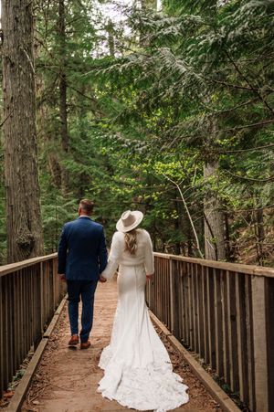 Bride and groom walking hand in hand on wooden bridge through lush green forest.