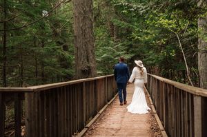 Bride and groom walk hand in hand down wooden boardwalk path through lush green forest.