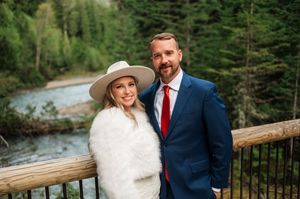 Bride in white fur wrap and wide-brim hat poses with groom in blue suit and red tie on wooden bridge.