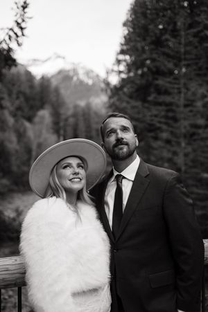 Black and white photo of a smiling bride in a wide-brim hat and fur wrap with her groom in a suit, mountains behind.