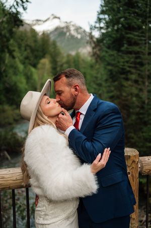 Groom in blue suit and red tie kisses bride in white fur wrap on mountain bridge outdoors.