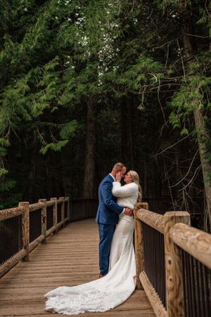 Bride and groom share a romantic kiss on a rustic wooden bridge surrounded by tall evergreen trees.