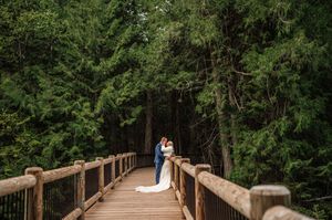 Bride and groom share a romantic kiss on a wooden bridge surrounded by tall evergreen trees.