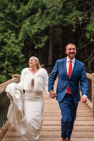 Happy bride and groom walk hand in hand on wooden bridge, woman in white dress fur shawl, man in blue suit red tie.