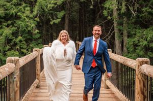 Smiling couple in wedding attire walking across wooden bridge surrounded by pine trees.
