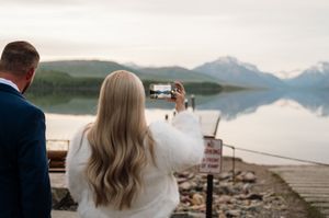 Bride in a white fur wrap photographs a serene mountain lake reflection from a dock during her wedding day.