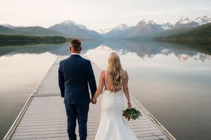 Bride and groom walking hand-in-hand down wooden dock toward stunning glacier lake and snow-capped mountain panorama.