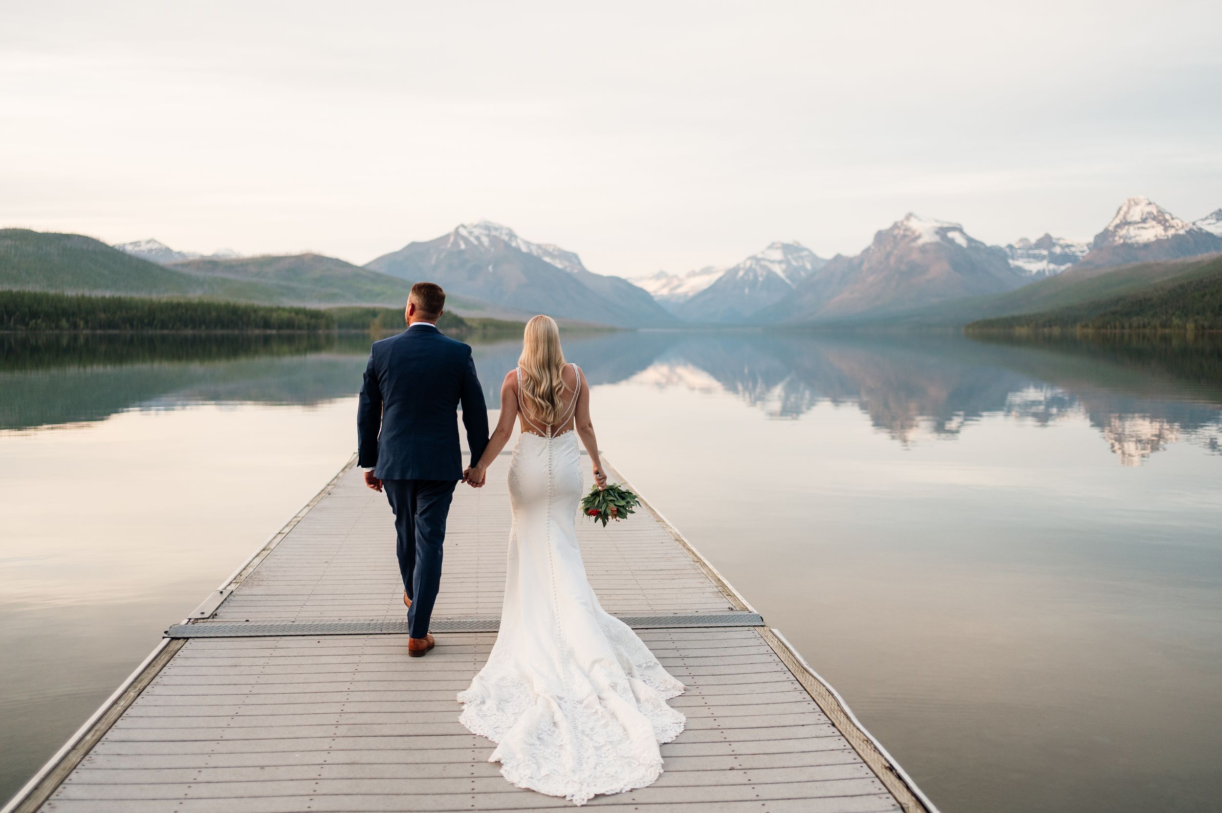 Bride and groom walk hand-in-hand down a dock toward stunning snow-capped mountains reflected in a calm lake.