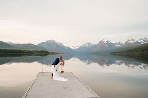 Couple shares a romantic dip kiss on a dock with stunning mountain reflections on a calm lake.