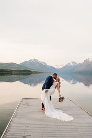 Groom dipping bride on dock with stunning mountain lake reflection in background.