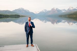 Groom in a navy suit adjusts his red tie on a dock with breathtaking snow-capped mountain lake views behind him.