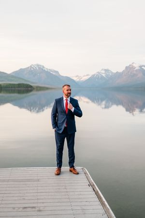 Groom in navy suit and red tie stands confidently on dock before stunning reflective mountain lake.