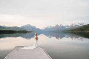 Bride in a flowing white gown stands alone on a dock surrounded by majestic mountains reflecting in a glassy lake.