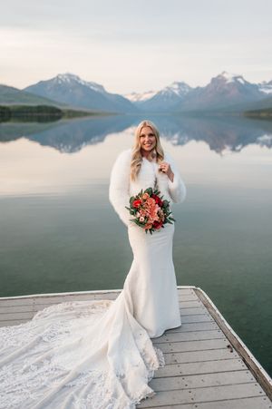 Bride in white gown and fur wrap holding bouquet on dock with mountain lake view.