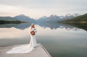 Bride in fitted lace gown holds bouquet on dock with mirror-like mountain lake and snow-capped peaks at sunset.