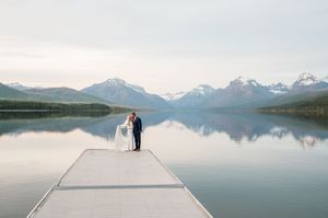 Bride and groom stand on a dock at a serene mountain lake with snow-capped peaks reflected in calm water.