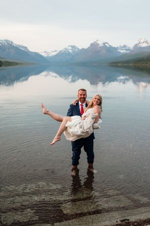 Groom carries bride while standing in mountain lake with stunning snowy peaks reflected in water.