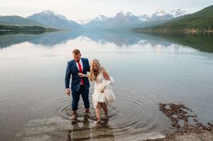 Bride and groom wade through scenic mountain lake with snow-capped peaks reflecting in calm water.