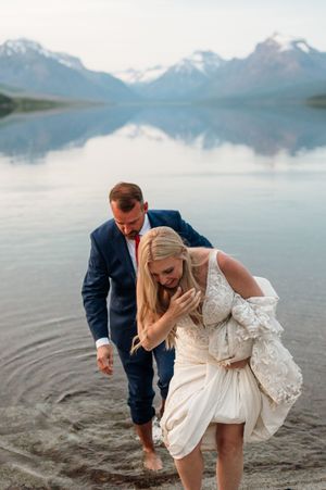 Couple wading barefoot in mountain lake, bride lifting her wedding gown from the water.