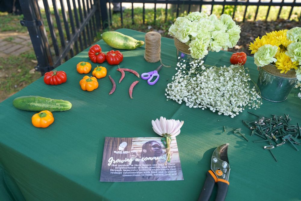 Outdoor table with fresh vegetables, flowers, gardening tools, and a program booklet on a green tablecloth.