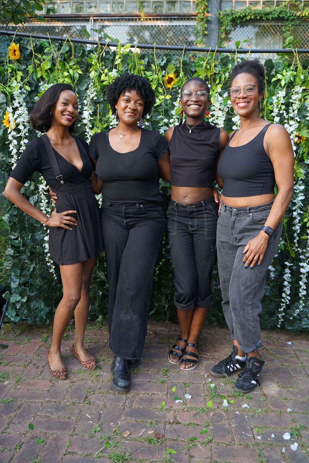 Four women in black outfits smiling together in front of a green floral wall backdrop outdoors.
