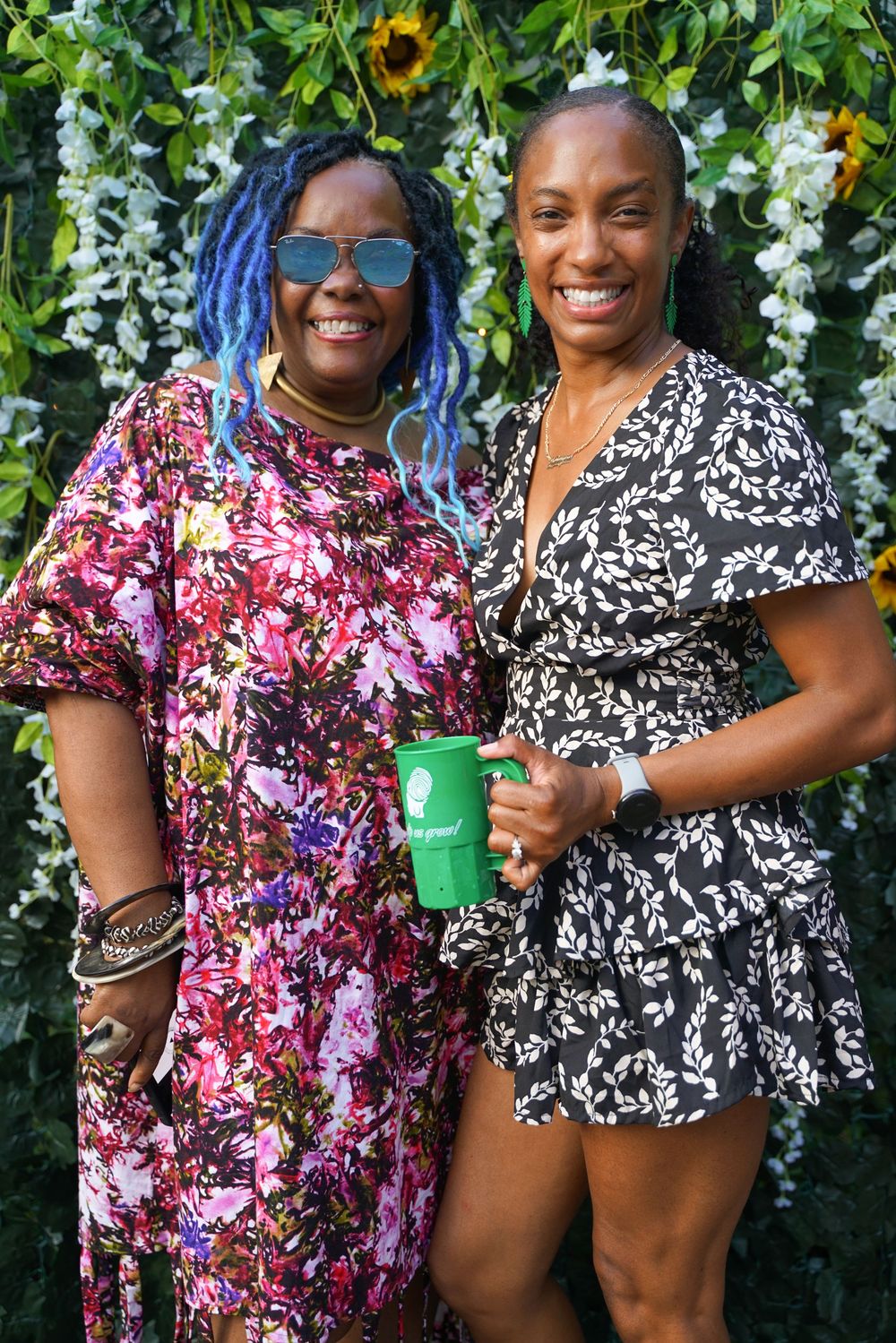 Two women smiling and holding green mugs in front of a floral backdrop with sunflowers.
