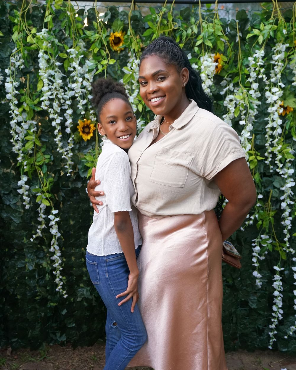 A smiling woman and young girl posing together in front of a floral backdrop with sunflowers.