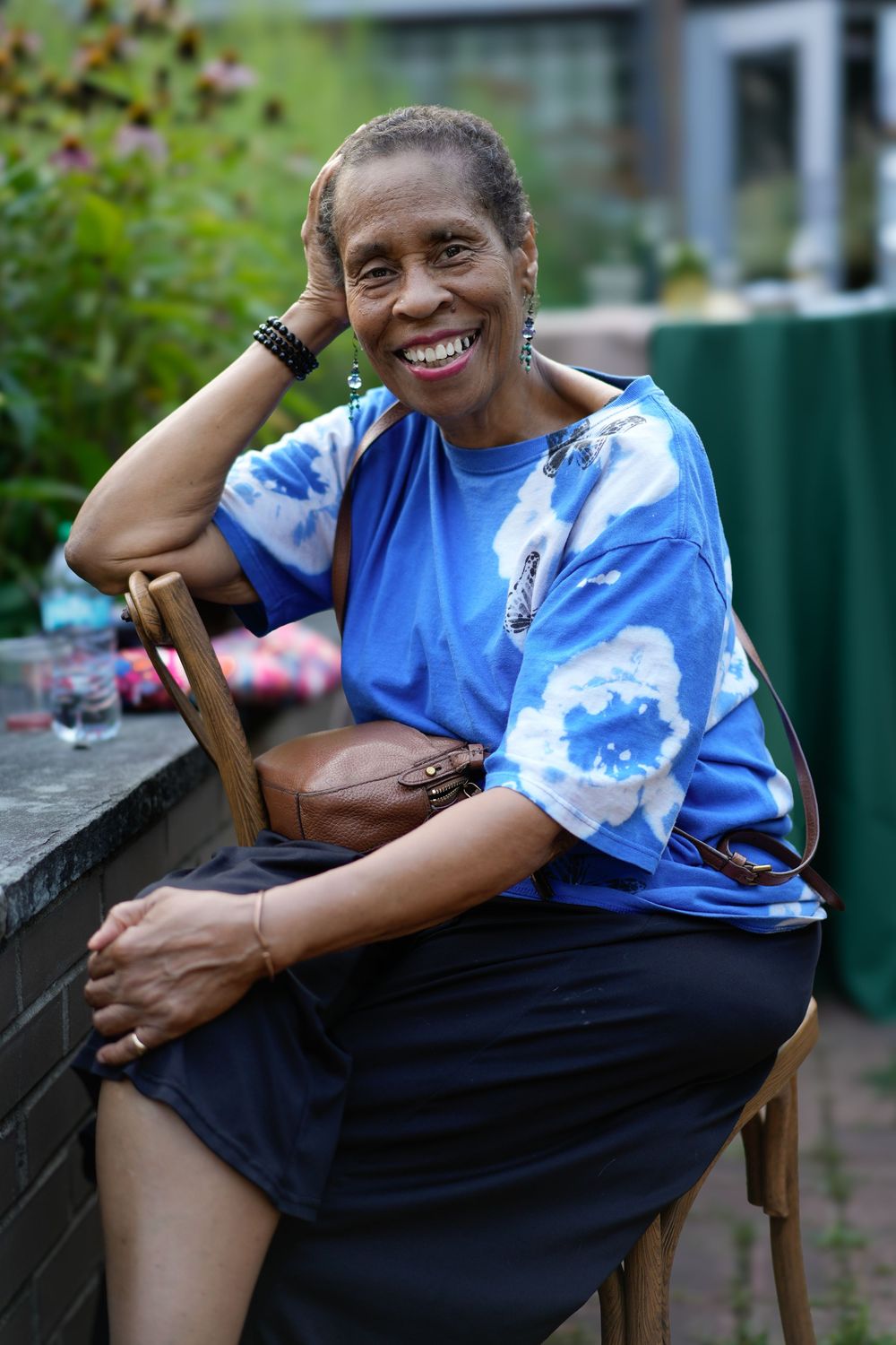 Smiling elderly Black woman in blue tie-dye shirt sits outdoors, holding a brown purse, with greenery in background.