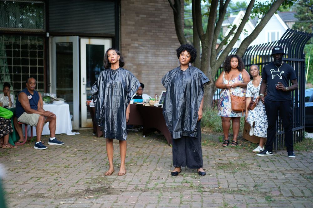 Two graduates in black gowns stand proudly outdoors during a graduation celebration with family and friends gathered nearby.