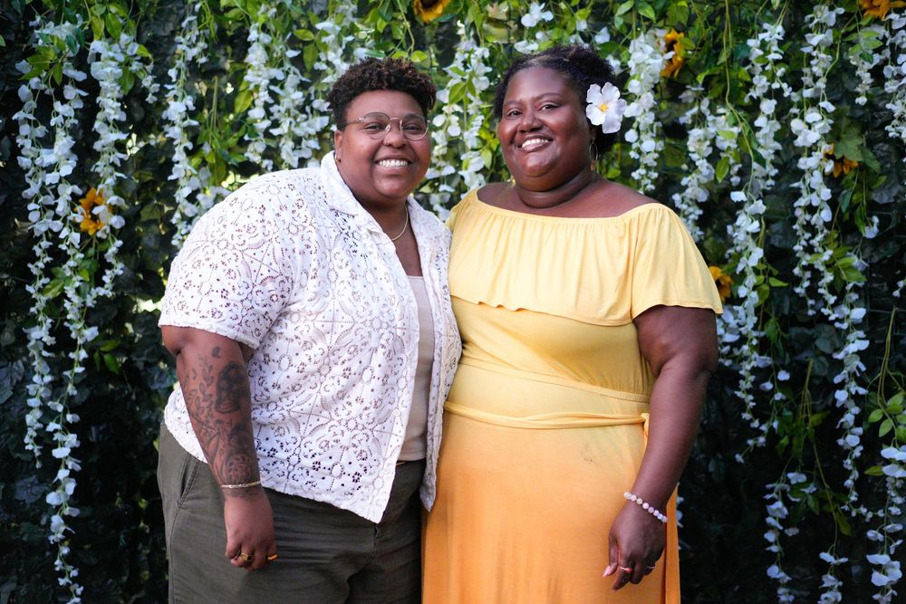 Two women smiling together in front of a green floral wall, one in yellow off-shoulder dress.