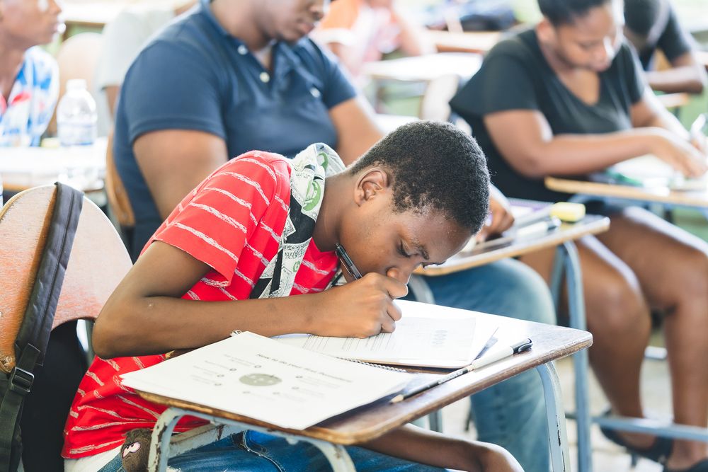 A young student in a red shirt focuses intently while writing at a school desk during an exam.