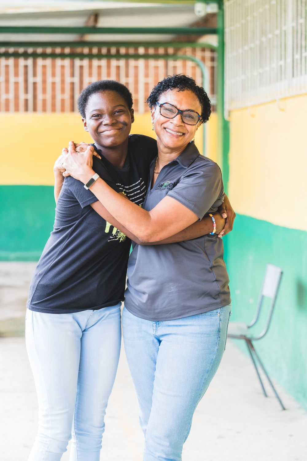 Two smiling women embracing each other outdoors near a colorful yellow and green wall.