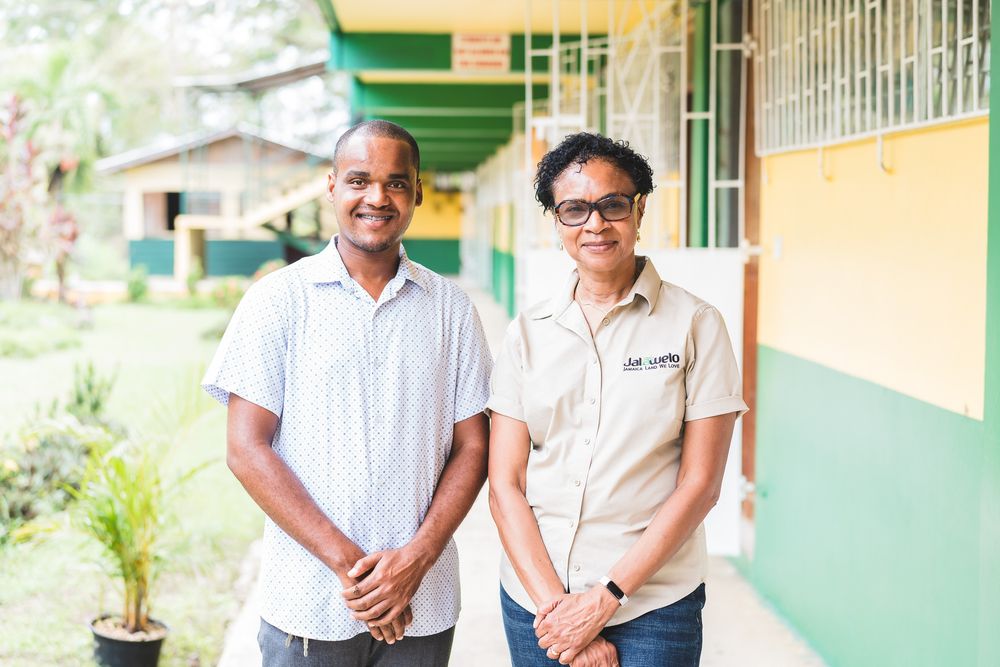 Two educators standing together outside a green and yellow school building in a tropical setting.
