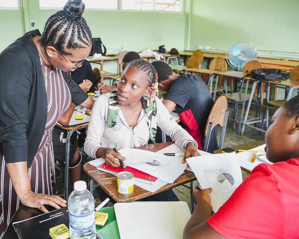A teacher in a black dress assists two students working at desks in a bright classroom.