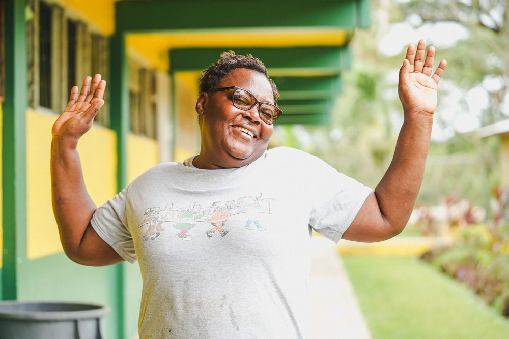 Joyful woman with glasses raising both hands outdoors near colorful building.