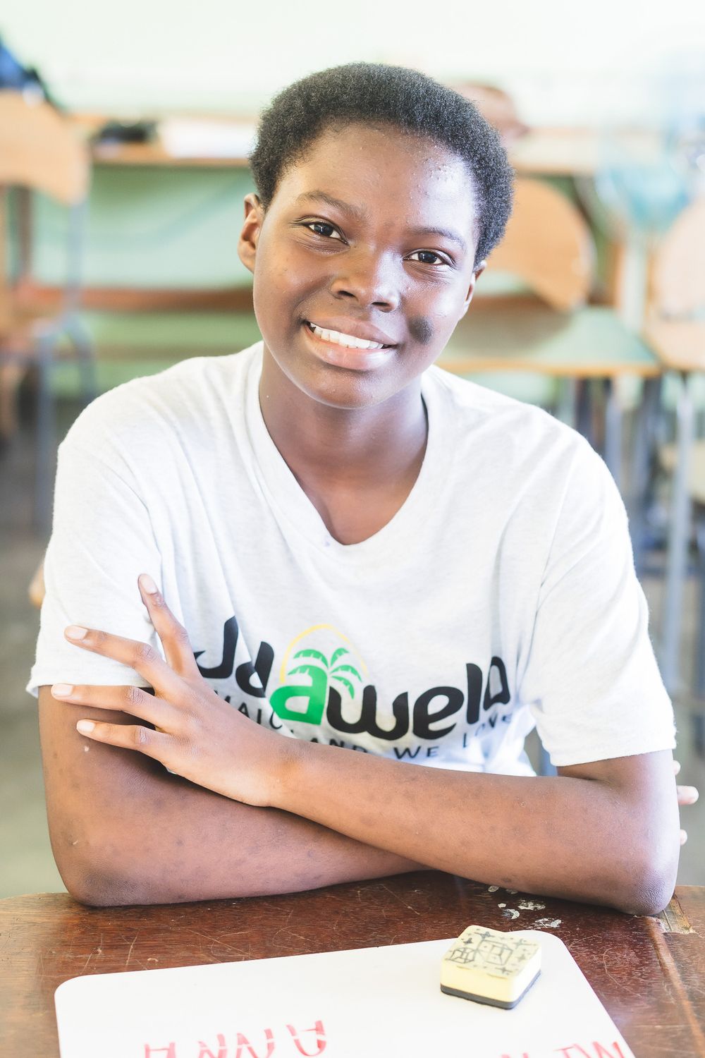 Smiling young African student in a white t-shirt sitting with arms crossed at a school desk.