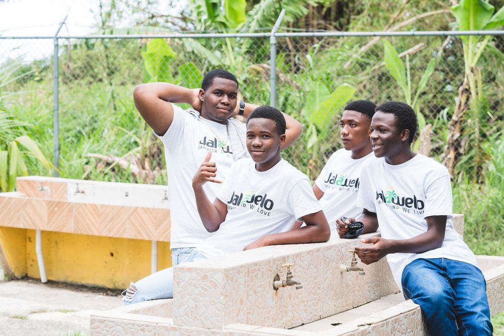 Four young men in matching white Jalebo t-shirts sitting outdoors near water taps in a lush green setting.