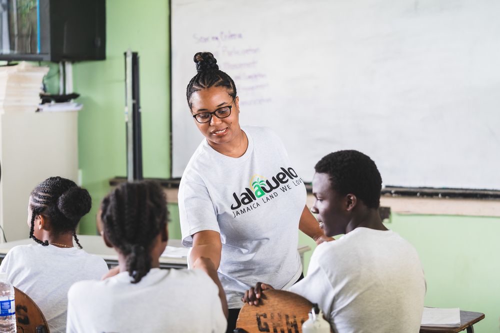 A teacher in a Jalguete shirt engages with students in a bright classroom with a whiteboard.