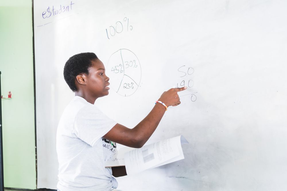 A young Black student writes math problems on a whiteboard during a classroom lesson.