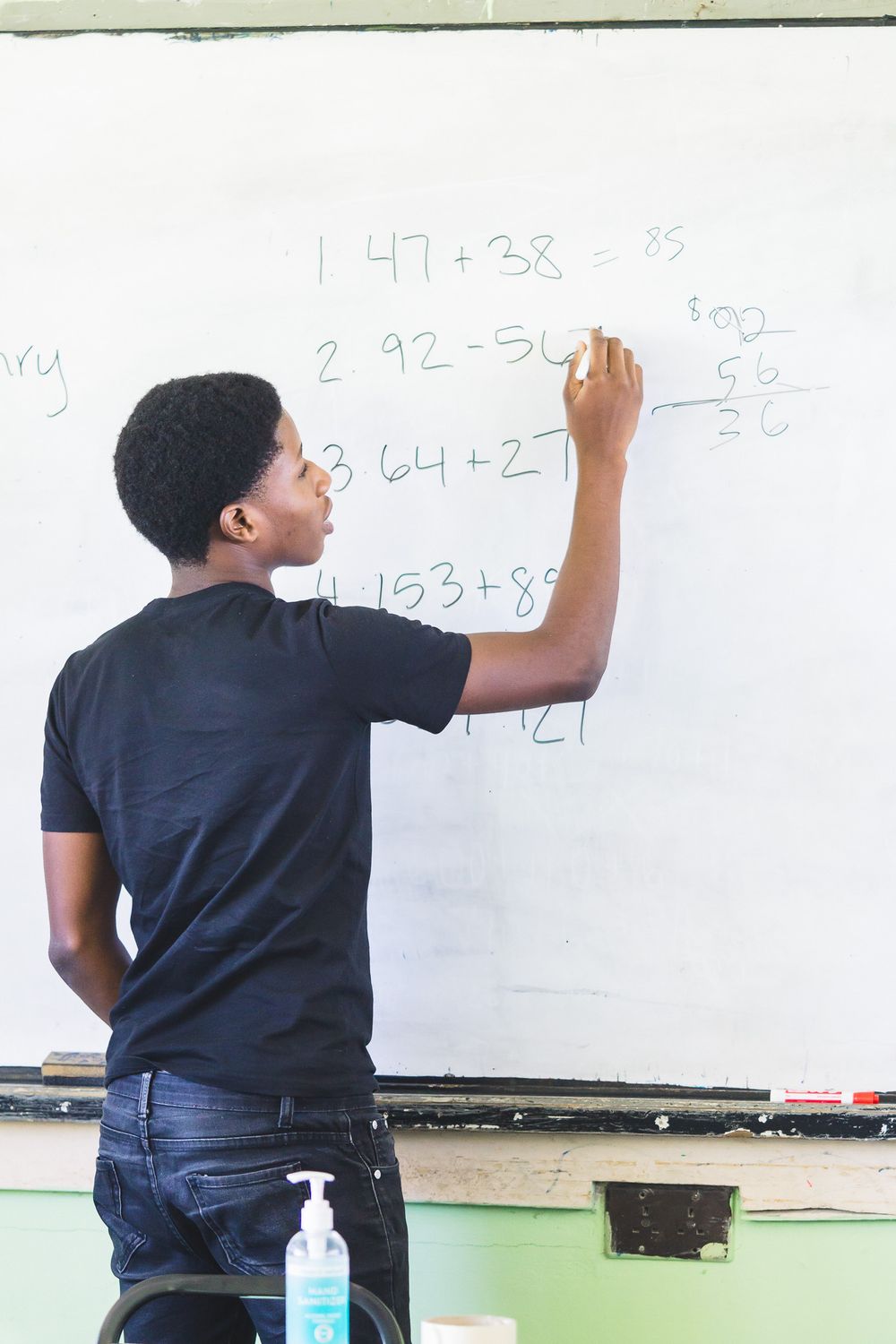 A student in a black shirt solves math problems on a whiteboard in a bright classroom.