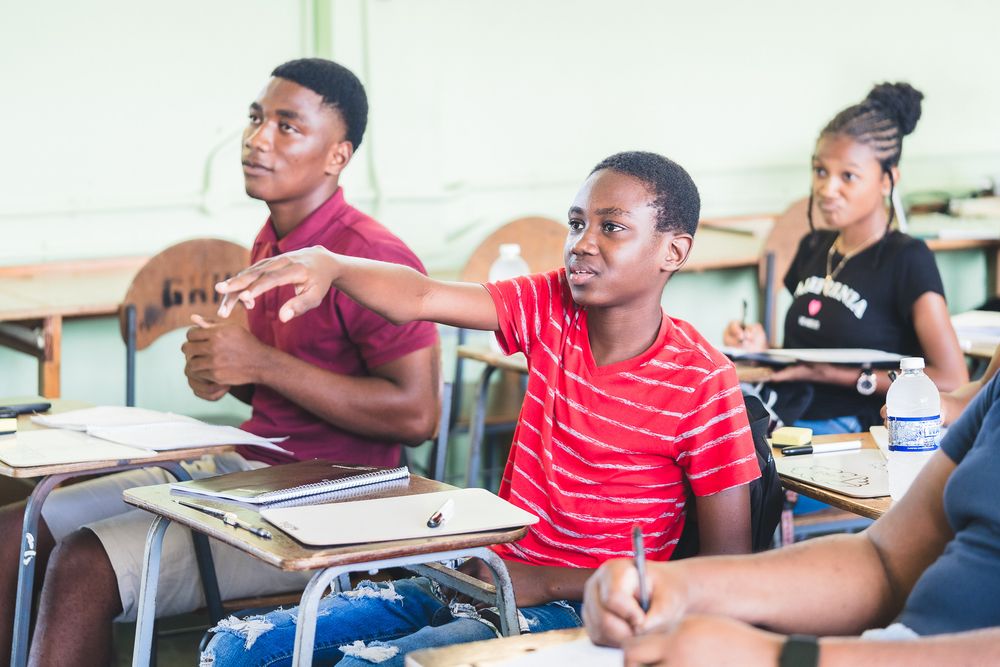 Students engage in classroom discussion at their desks, with a boy in a red shirt gesturing expressively.