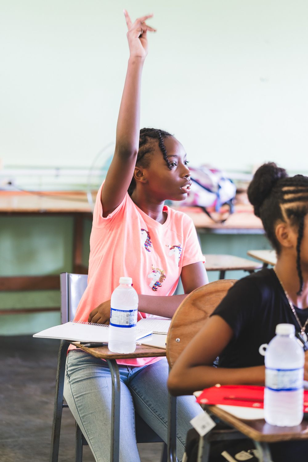 A young girl in a pink shirt raises her hand eagerly to answer a question in a classroom.