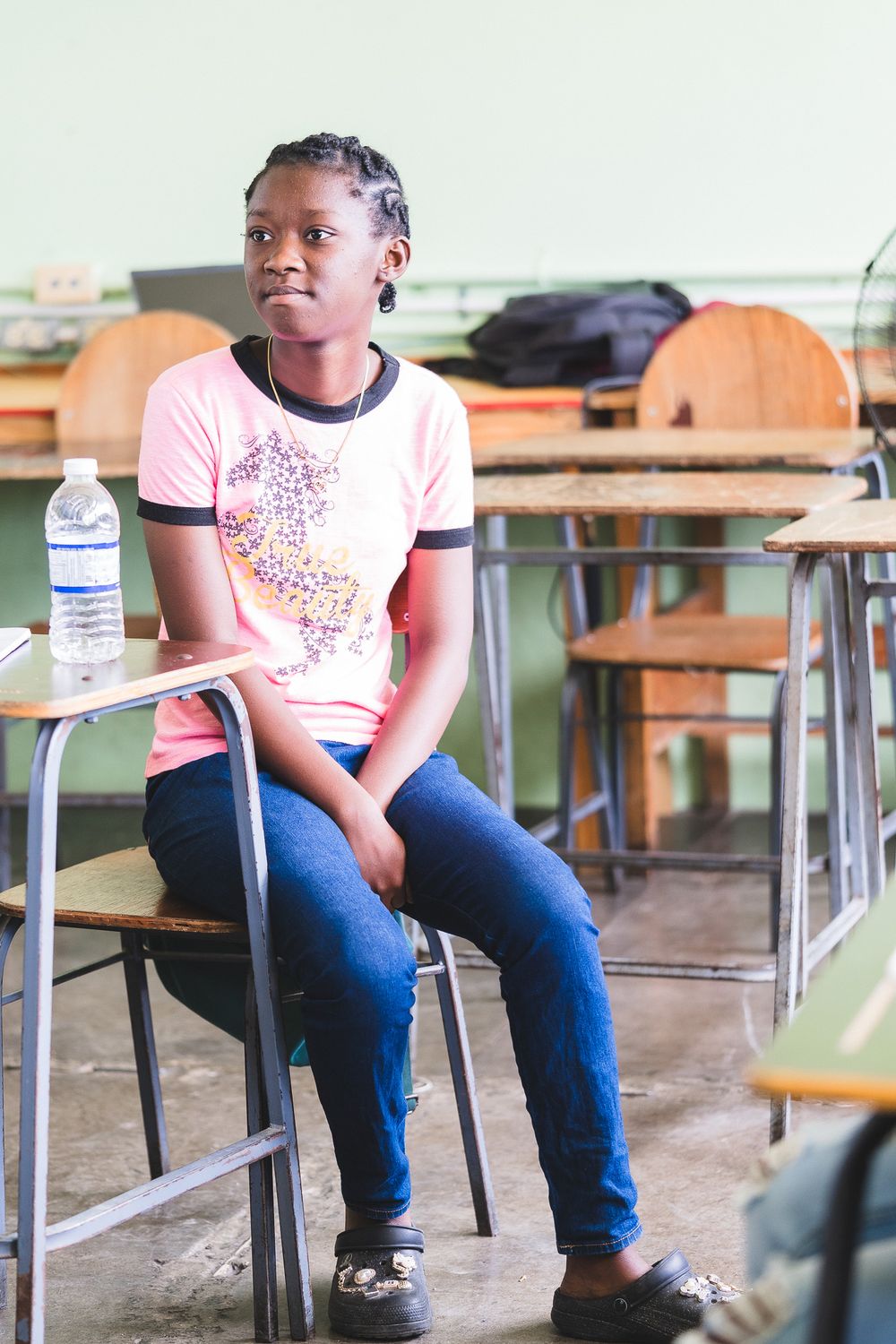 Young African girl in pink shirt sitting at a school desk with a water bottle in a classroom.